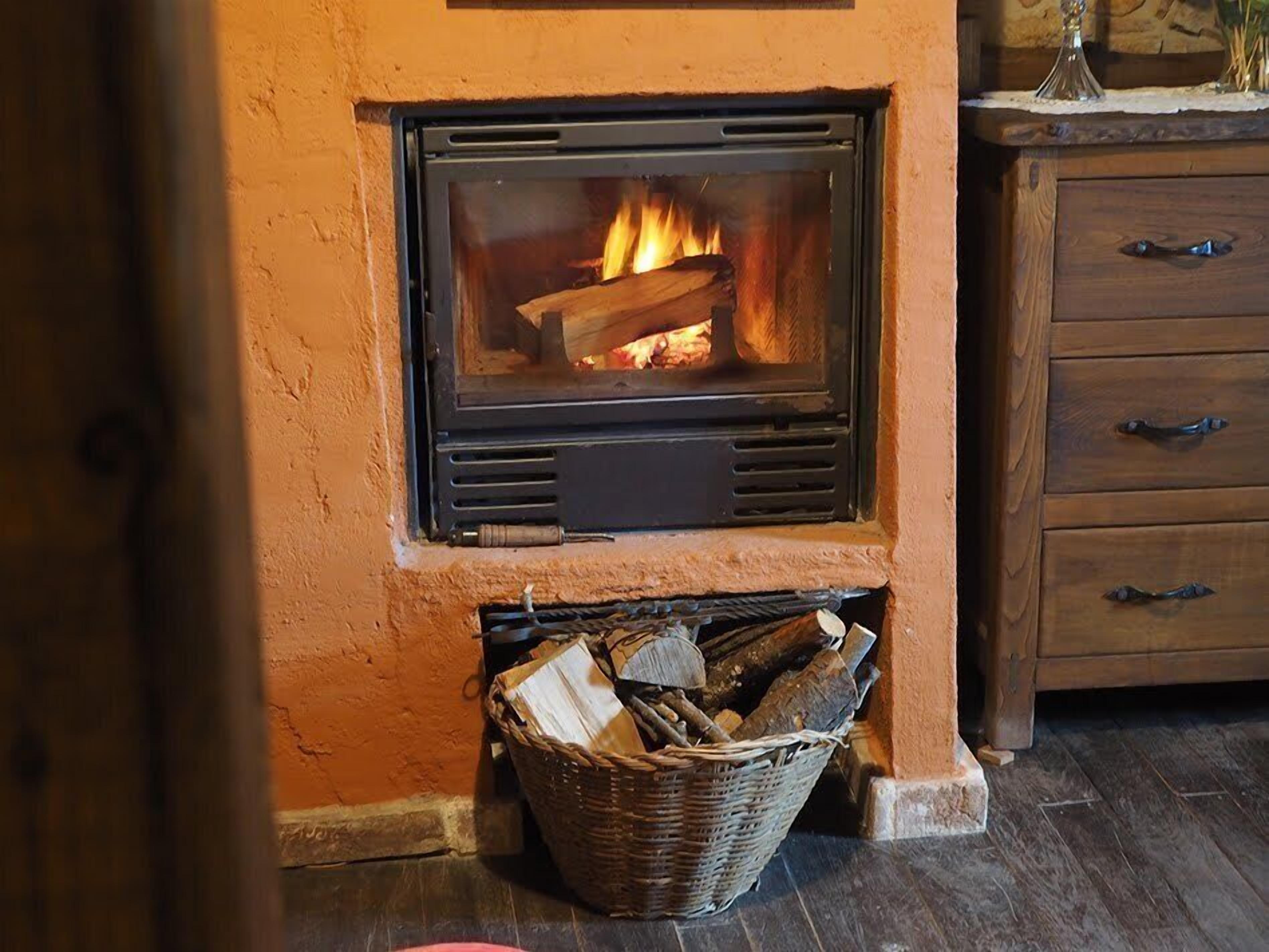 A fireplace with a fire burning, a wooden cabinet to the right, and a basket of firewood below.
