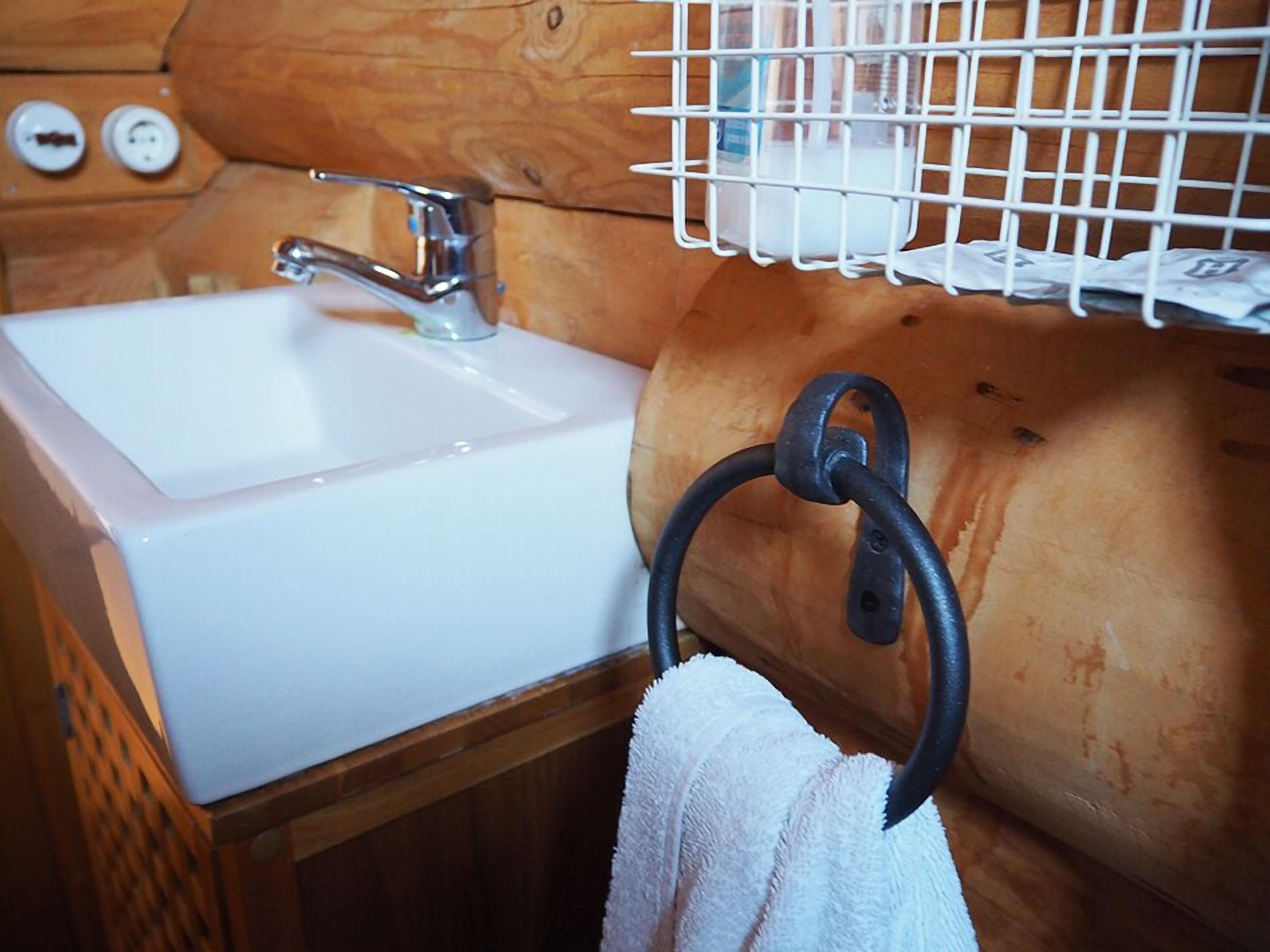 L’intérieur d’une cabane en bois, avec un évier blanc, une serviette suspendue à un anneau métallique et un récipient en plastique blanc posé sur une étagère.