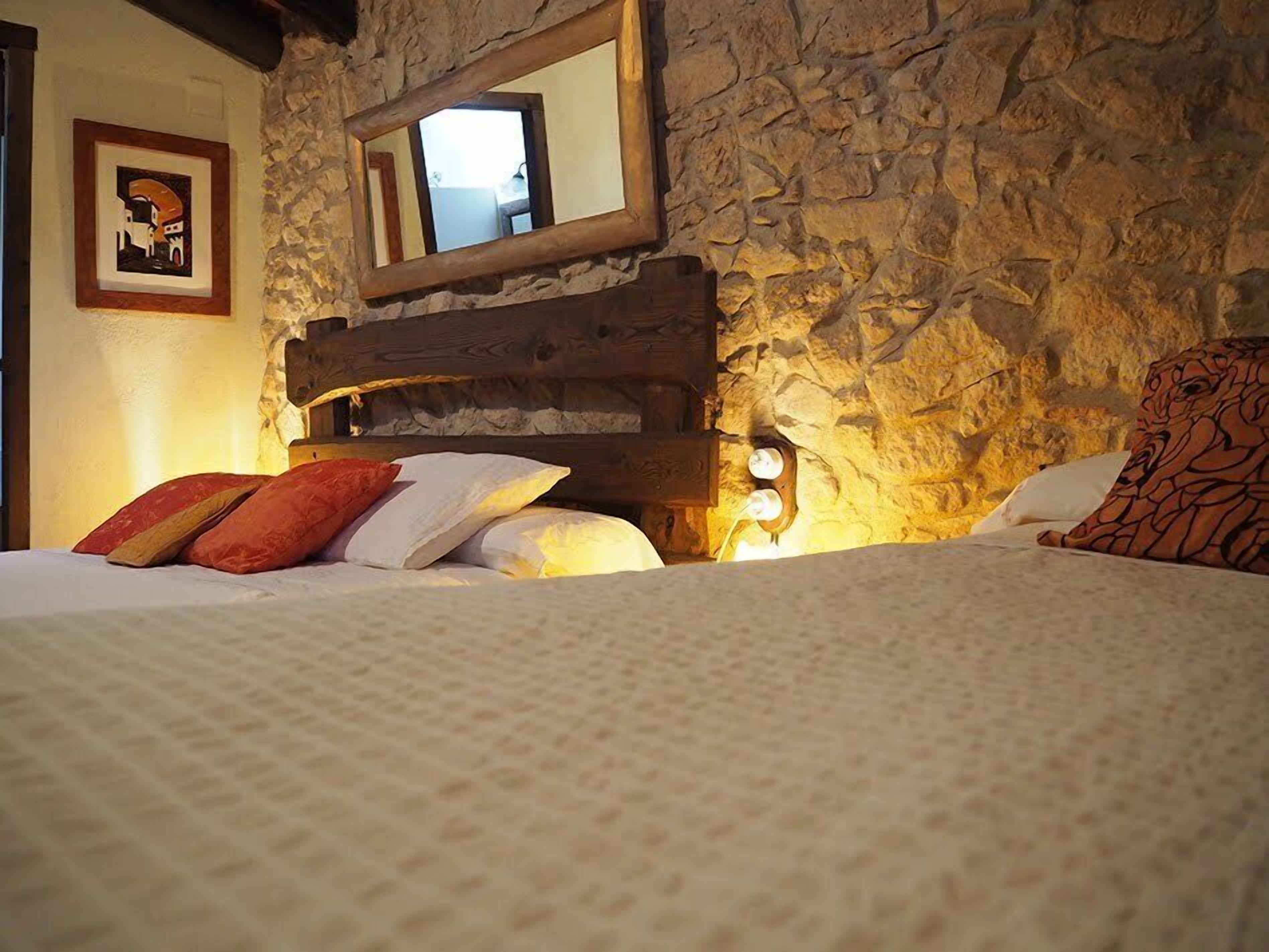 A bed with a stone wall and a wooden headboard, a framed picture on the wall, and a mirror above the headboard.