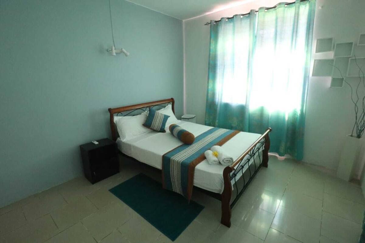 A bedroom with a bed, a bedside table, a nightstand, a window with curtains, and a wall-mounted shelf.