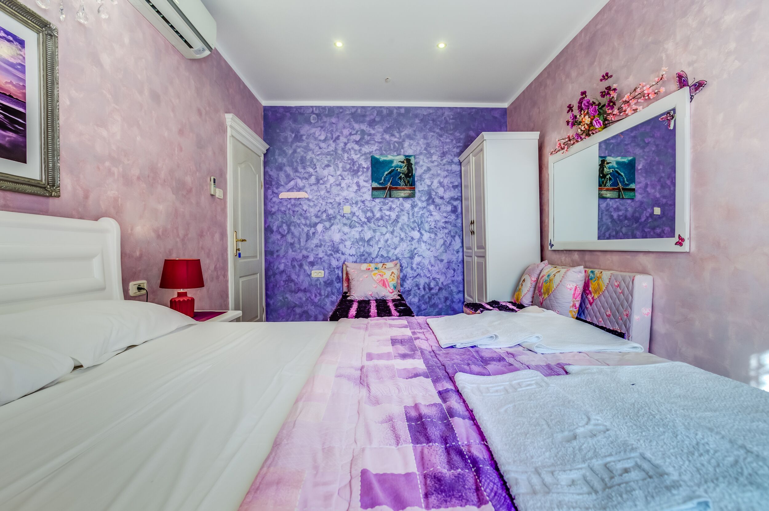 A bedroom with a bed, a nightstand, a red lamp, a mirror, and a door.
