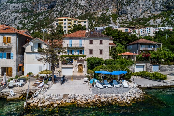 A coastal hotel with a stone facade, a central entrance, and a pool area with lounge chairs.