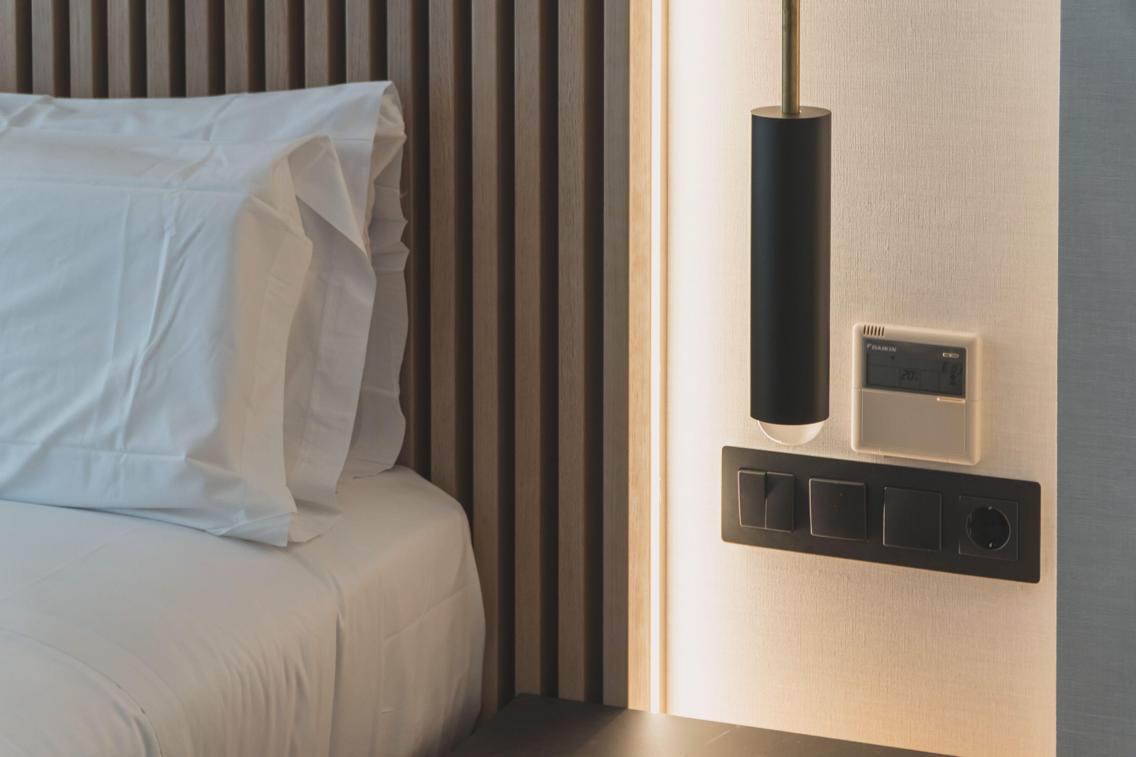 Une chambre d’hôtel moderne avec un lit soigneusement fait, un panneau de commande fixé au mur et un luminaire vertical.