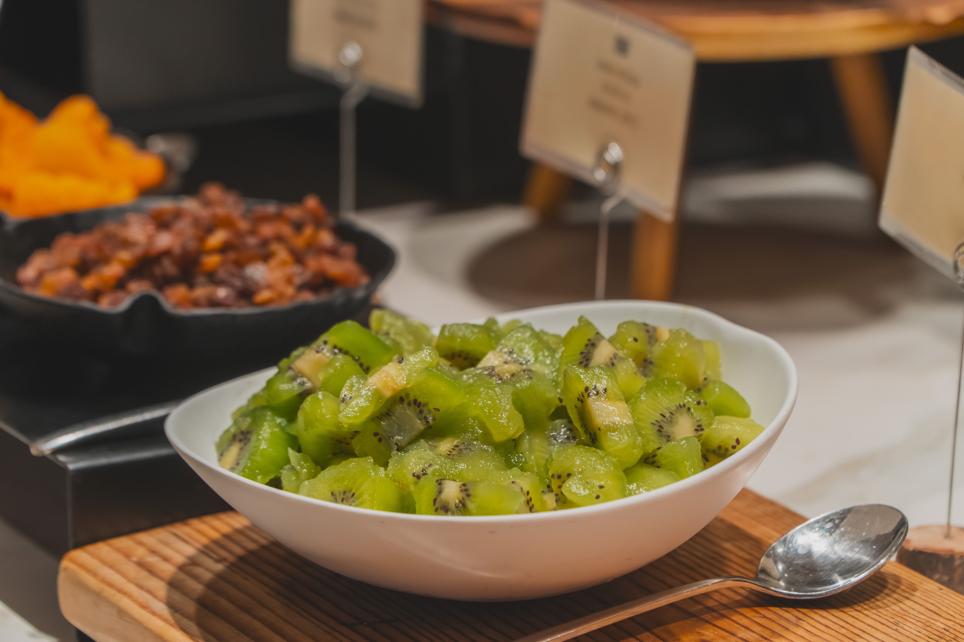 A bowl of sliced kiwi fruit on a wooden board with a spoon beside it.