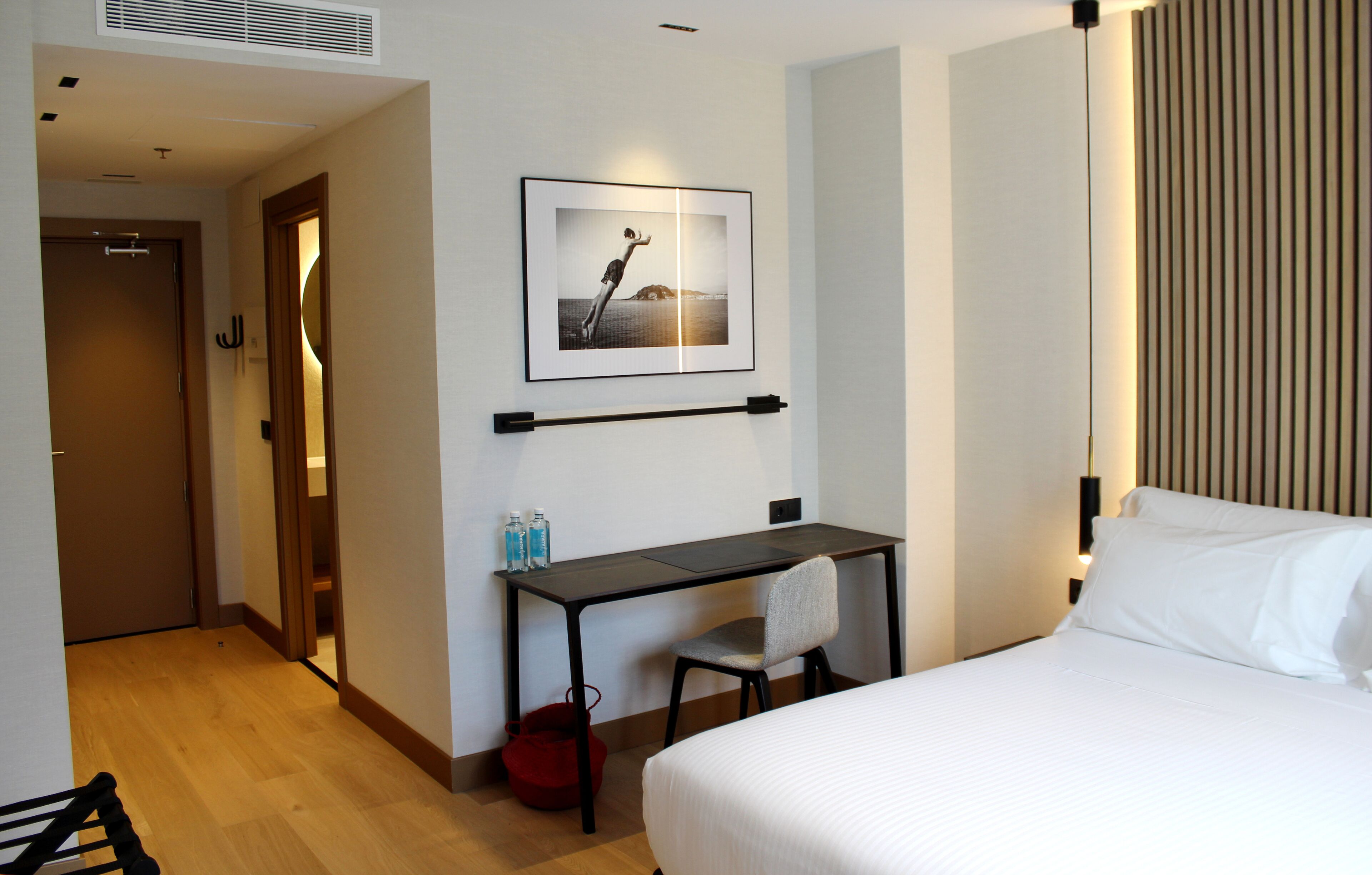 Habitación de hotel con cama, escritorio, silla, un cuadro en la pared y una bolsa roja en el suelo.
