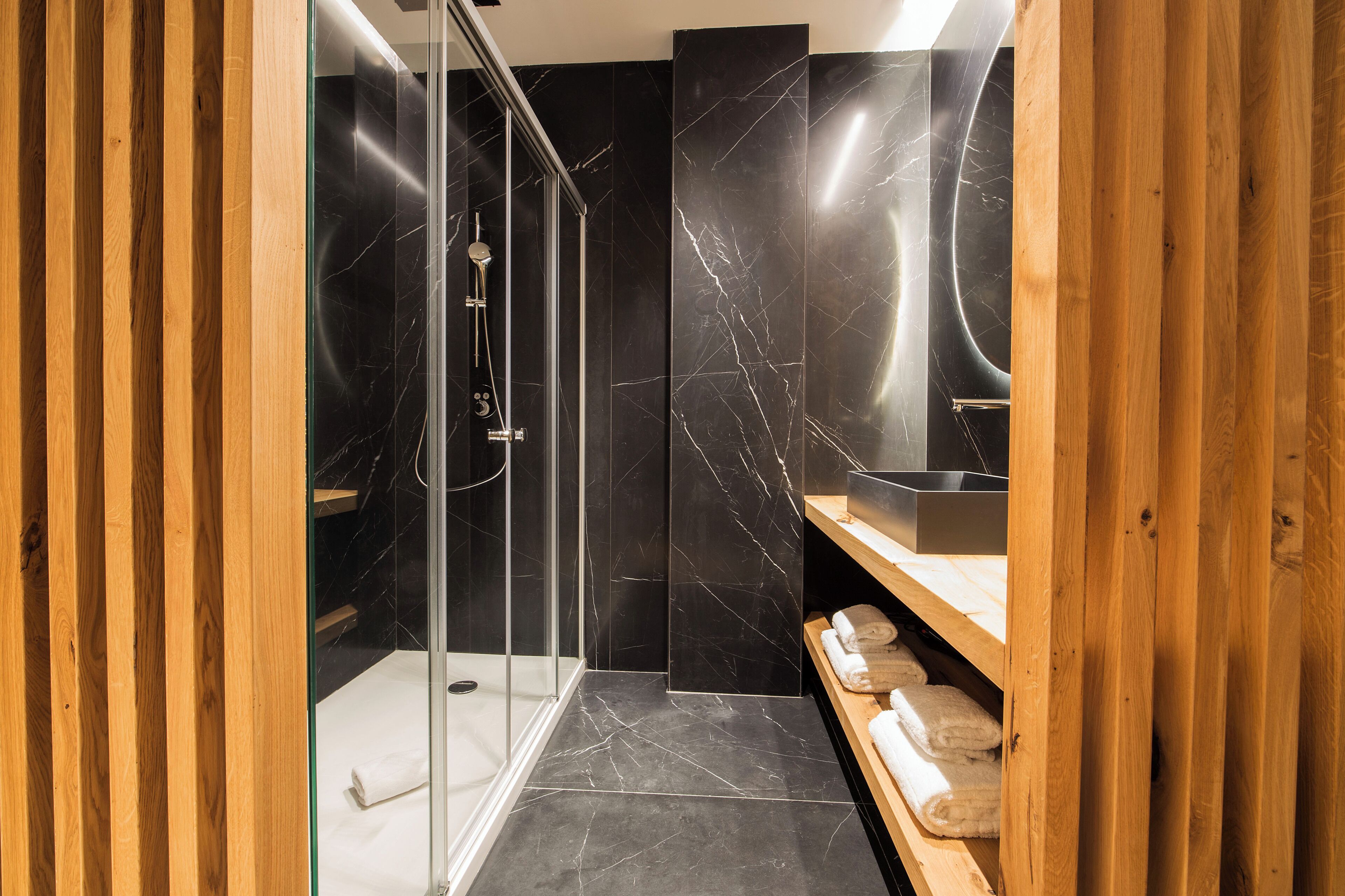A modern bathroom with a glass-enclosed shower, marble walls, wooden shelving with towels, and a sleek sink.
