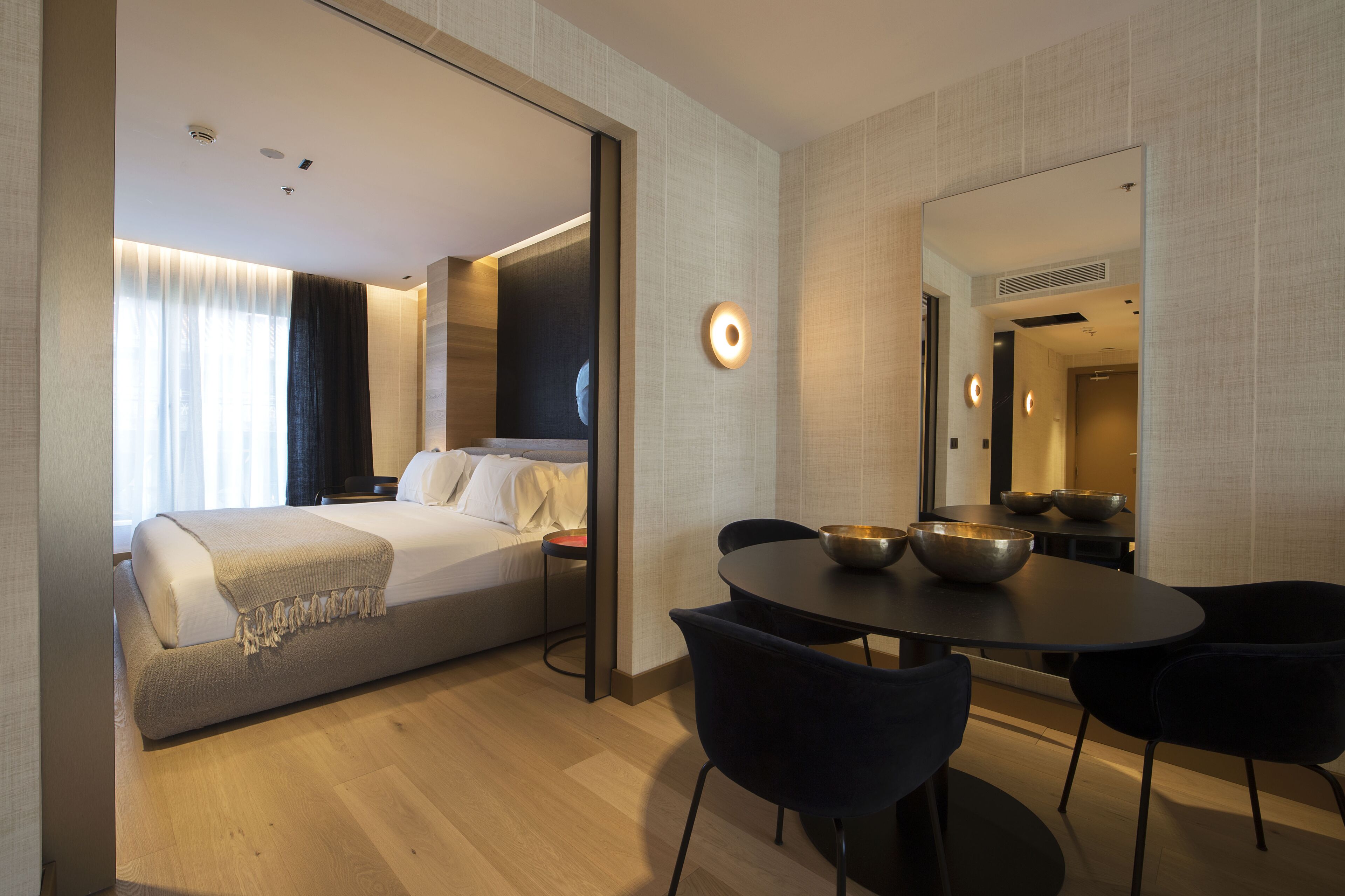 A modern hotel room with a large bed, a dining table with bowls, and a mirror reflecting the room.