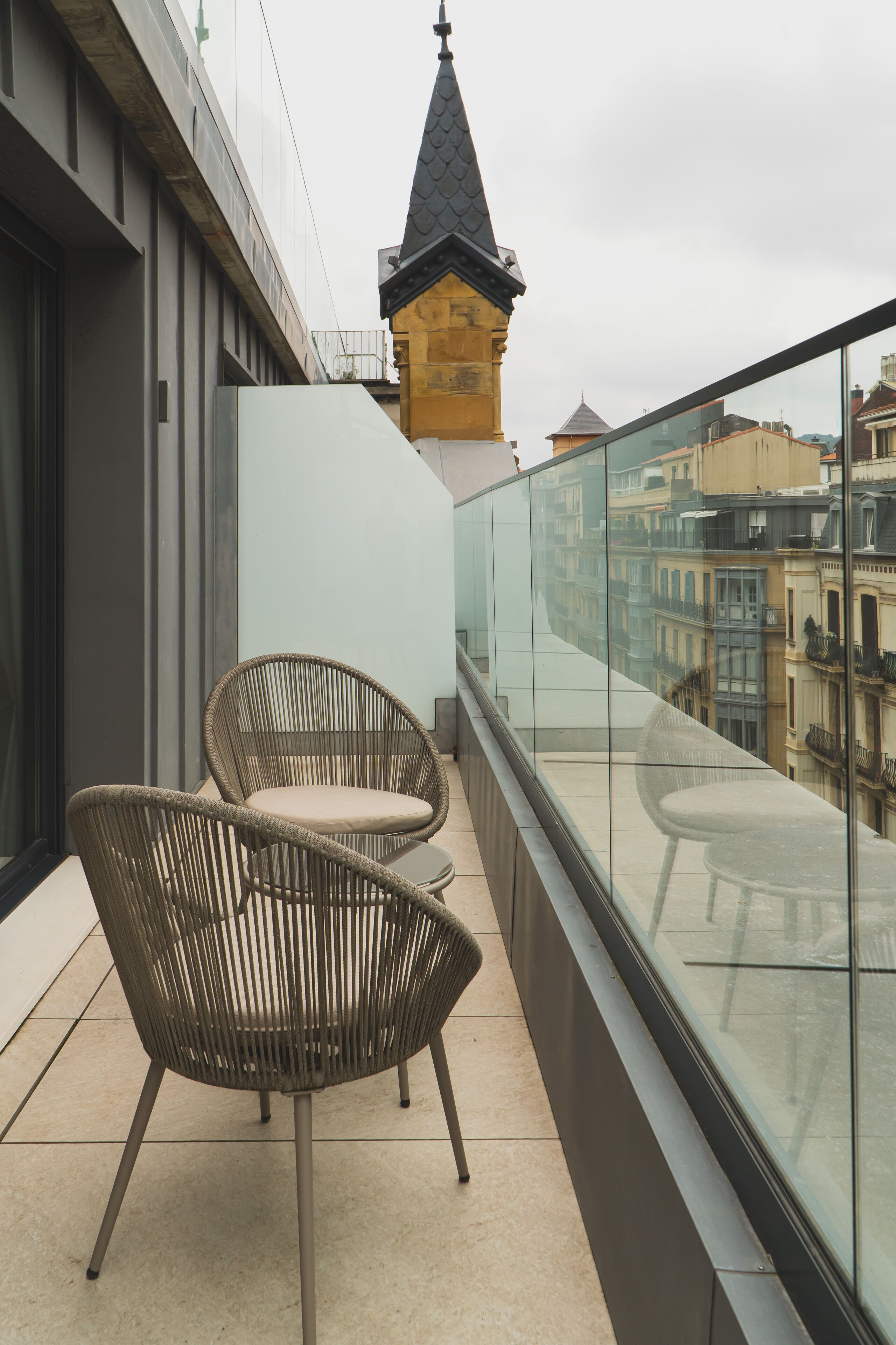 A balcony with two wicker chairs, a glass railing, and a view of historic buildings.