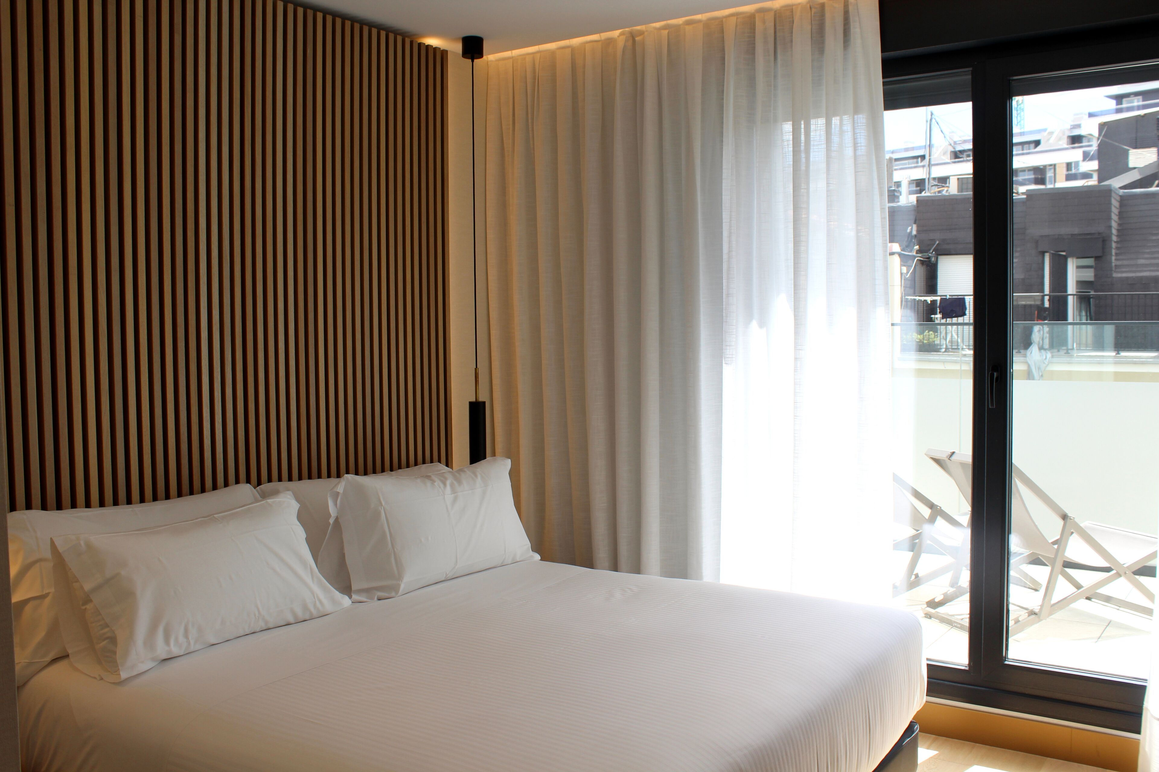 Habitació d'hotel moderna amb un llit gran, un panell de capçalera de fusta i vista a la ciutat a través d'una finestra gran.