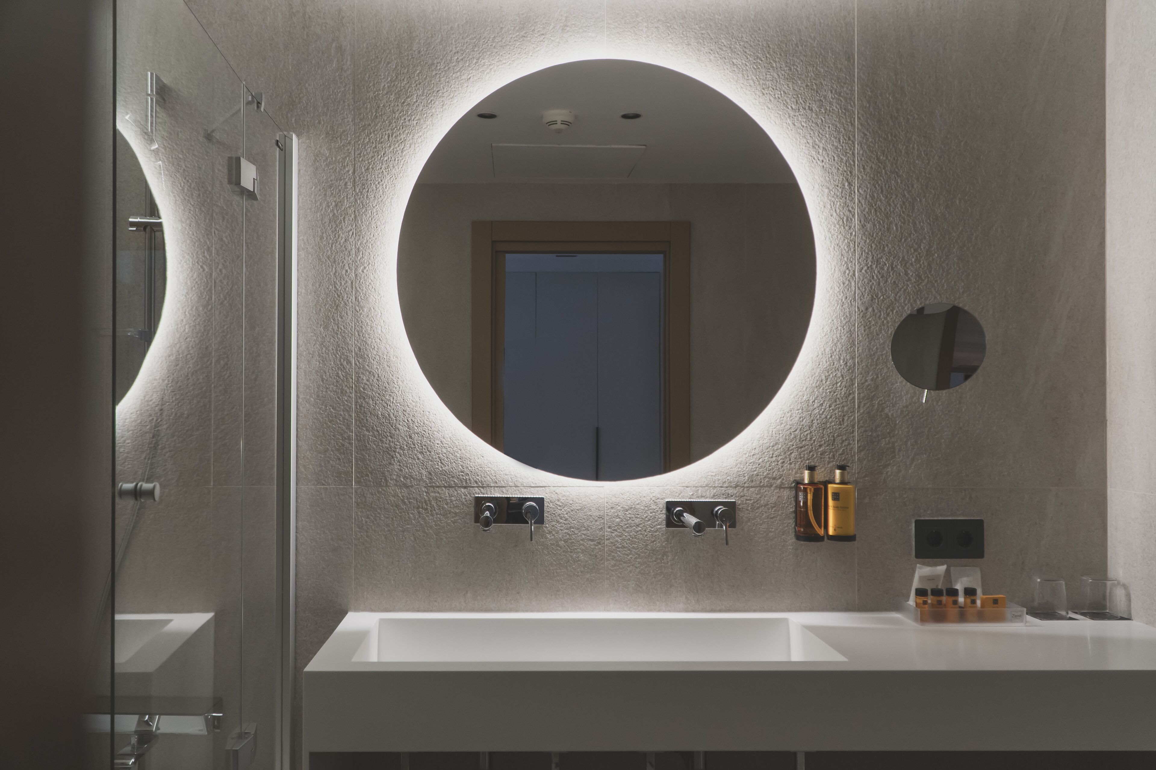 A modern bathroom with a large round mirror, a white sink, and a glass shower enclosure.