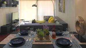 A dining area with a table set for two, featuring black plates, silverware, and a bottle of wine. A sofa with cushions is visible in the background, and a decorative wall piece is on the right. - Metro Court Apartments (Limassol)
