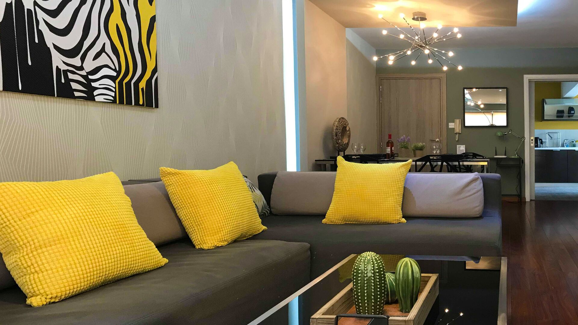 Una sala moderna con un sofá gris, cojines amarillos, una mesa de centro con un cactus y un comedor al fondo.
