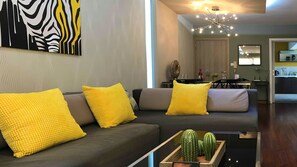 A modern living room with a grey sofa, yellow cushions, a glass coffee table with a cactus, and a dining area in the background.