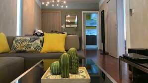 A modern living room with a sofa, a coffee table with decorative items, a ceiling light fixture, and a view into the kitchen. - Metro Court Apartments (Limassol)