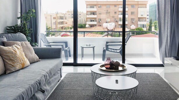 A modern living room with a grey sofa, a round coffee table with decorative items, and a view of a balcony with outdoor furniture and buildings in the background. - Metro Court Apartments (Limassol)