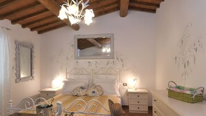 Une chambre avec un plafond en bois, un lit recouvert d’une literie à motifs floraux, des tables de chevet avec des lampes, un miroir et un panier vert posé sur une commode.