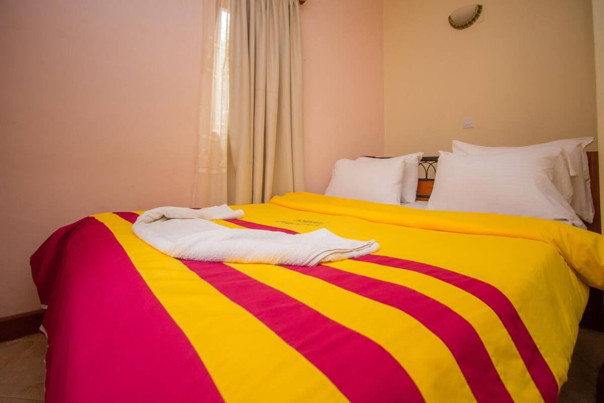 A hotel room with a bed featuring a striped yellow and red bedspread, white pillows, and a neatly folded white towel on the bed.