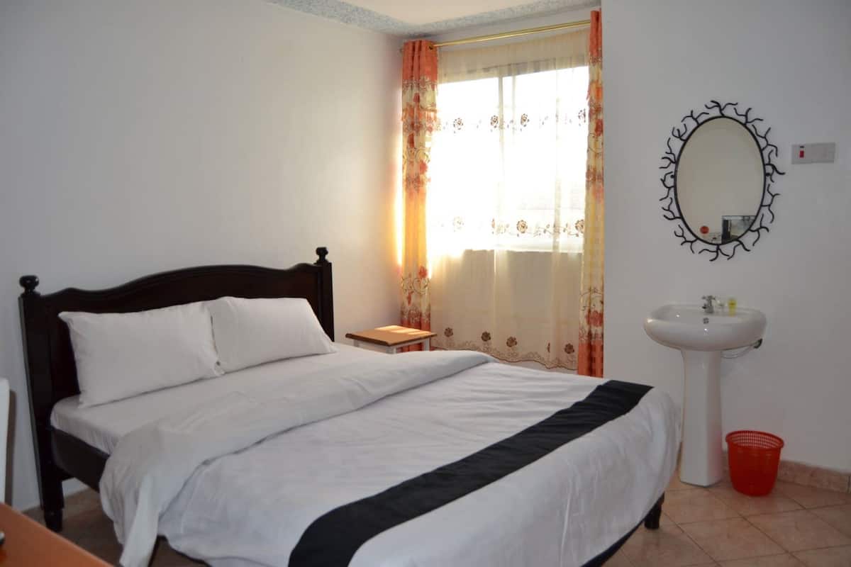 A bedroom with a bed, a bedside table, a window with curtains, a wall-mounted mirror, and a sink.