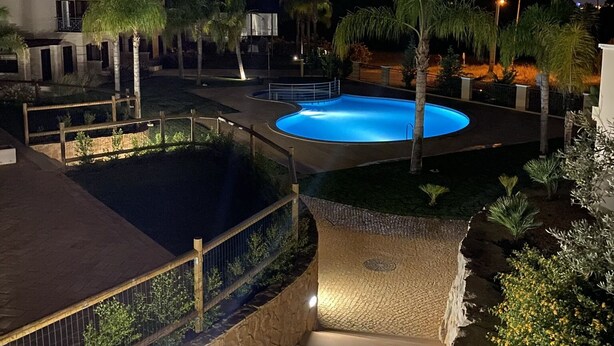 Vista nocturna d'una zona de piscina amb terrassa de pedra, palmeres i un edifici al fons.