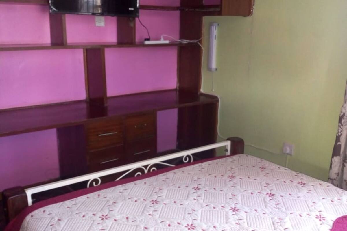 Un dormitorio con cama, televisor montado en una pared rosa y cómoda de madera.