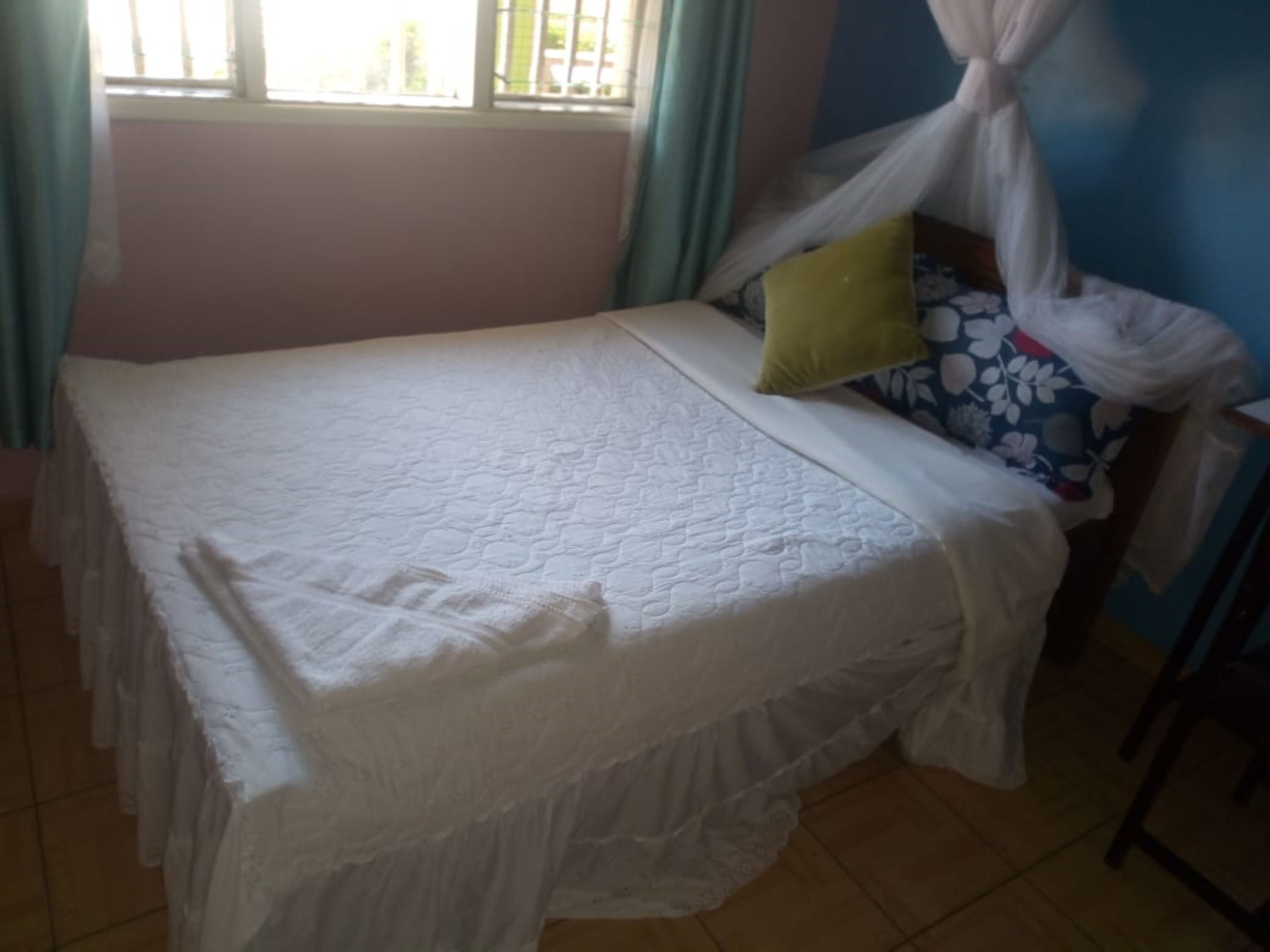 Una cama con una colcha blanca y dosel, un almohadón verde y un almohadón con motivos florales sobre un suelo de baldosas.