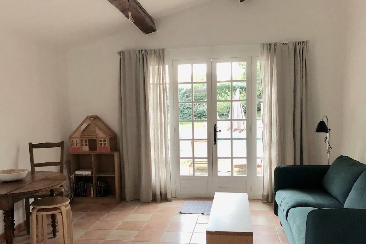 A living room with a sofa, a wooden table with a bowl, a small house model, and a door leading outside.