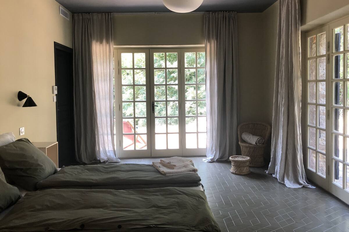 A bedroom with two beds, a large window with curtains, a wall-mounted lamp, and a wicker chair.