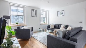 A modern living room with a grey sofa, a glass coffee table, and a white rug. There are two windows with blinds, a radiator, and a plant on the windowsill. - Fitzrovia Spacious 2BR2BA by Bloomsbury (London)
