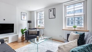 A modern living room with a flat-screen TV, comfortable seating, large windows, and minimalist decor. - Fitzrovia Spacious 2BR2BA by Bloomsbury (London)