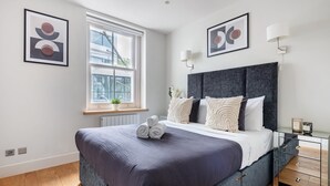 A modern bedroom with a large bed, a window, and artwork on the walls. - Fitzrovia Spacious 2BR2BA by Bloomsbury (London)
