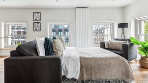 A spacious bedroom with a large bed, two seating areas, and multiple windows offering city views.
