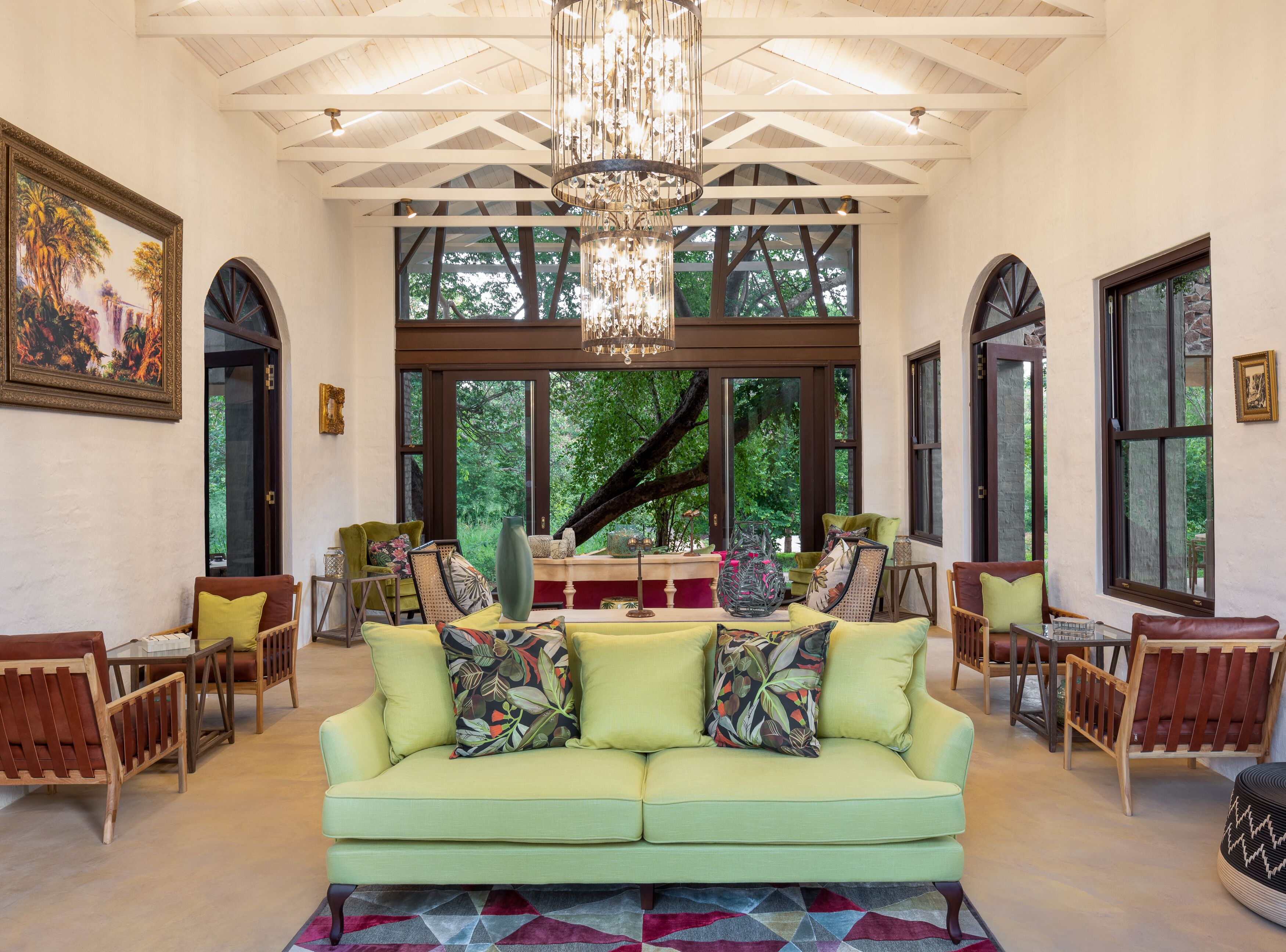 A spacious living room with a large green sofa, wooden chairs, and a chandelier. Large windows provide views of the outdoors.