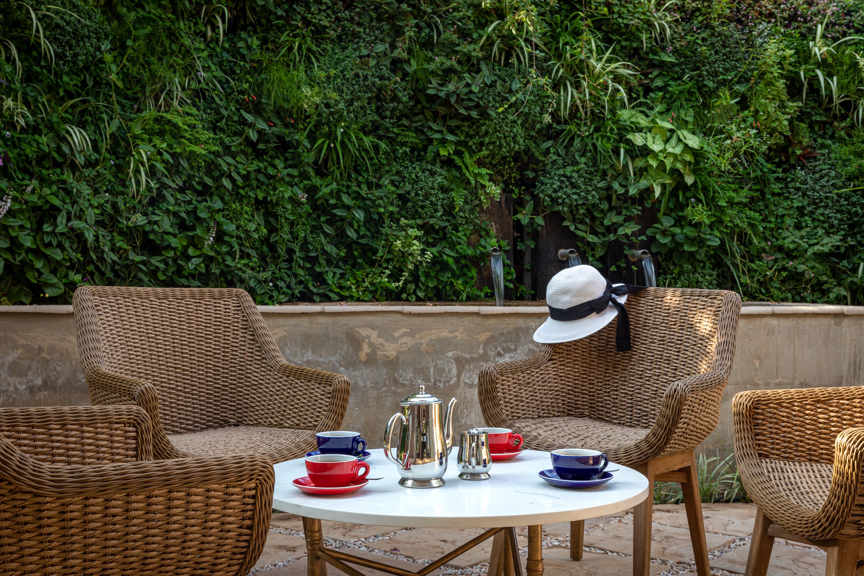 Des chaises en osier et une table avec une théière et des tasses, le tout placé devant un mur verdoyant.