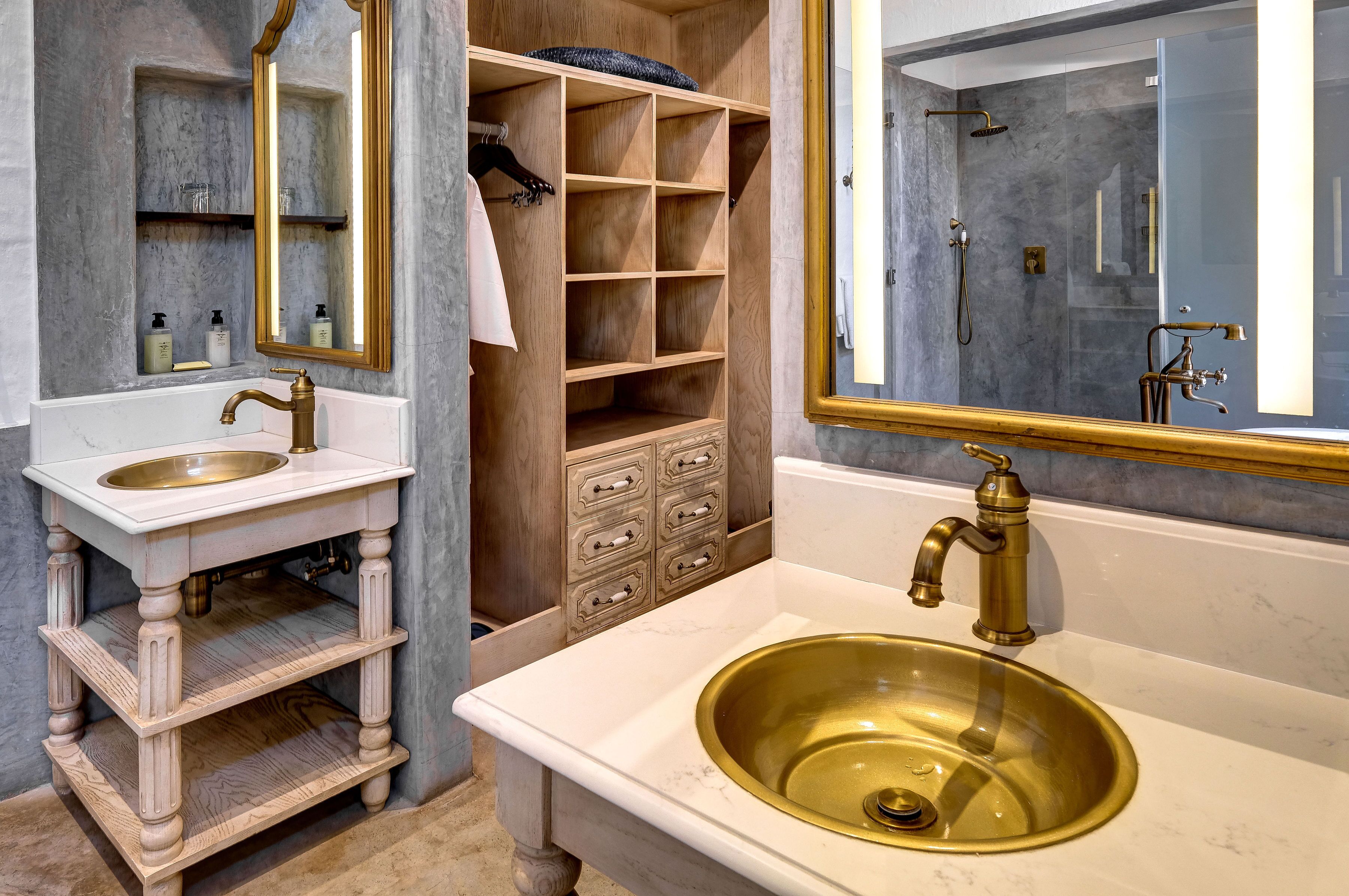 A bathroom with a white vanity, a gold sink, a wooden storage unit, and a glass shower enclosure.