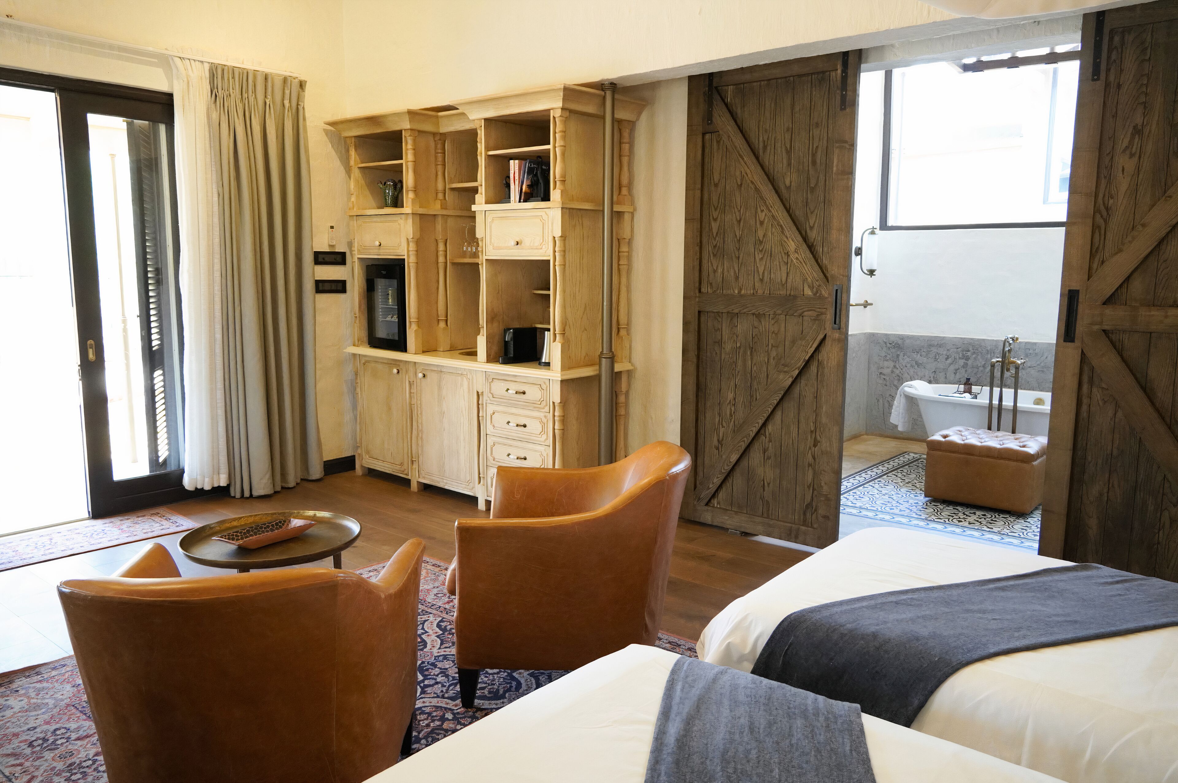 A hotel room with a bed, two chairs, a desk with a computer, a bookshelf, a wardrobe, and a view of the outside.