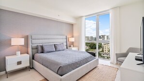 Un dormitorio moderno con una cama grande, dos mesitas de noche con lámparas, un televisor y vistas de la ciudad a través de amplios ventanales.