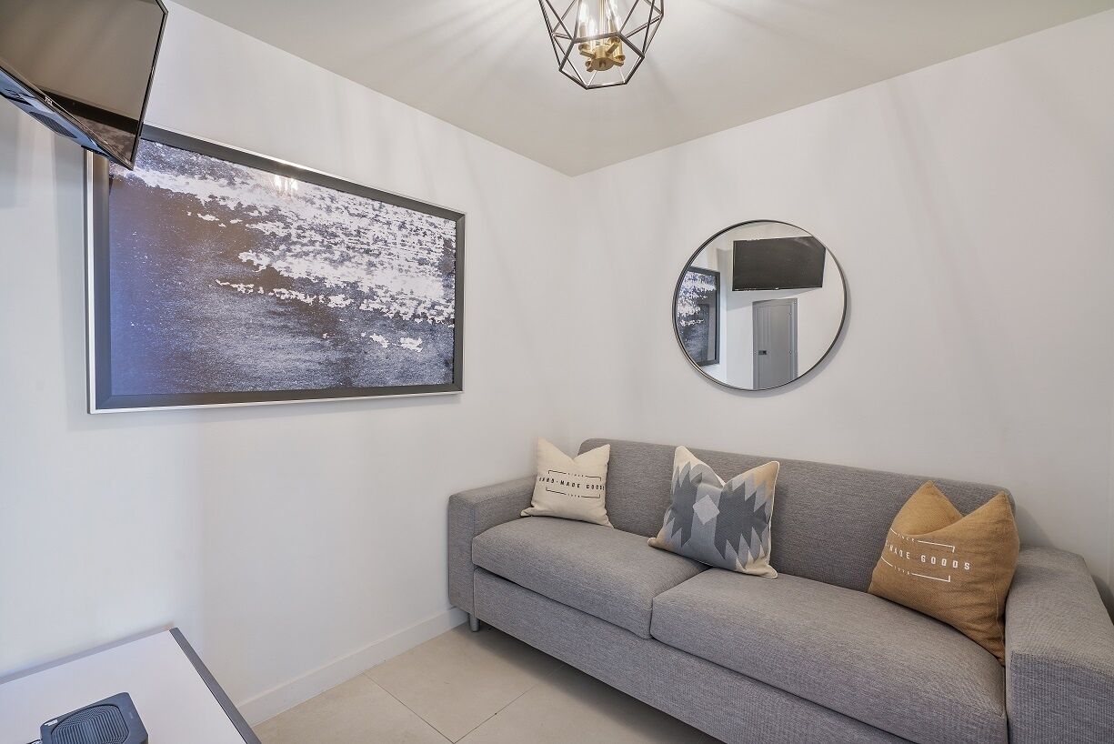 Un soggiorno moderno con un divano grigio, un quadro astratto incorniciato, uno specchio rotondo e un lampadario.