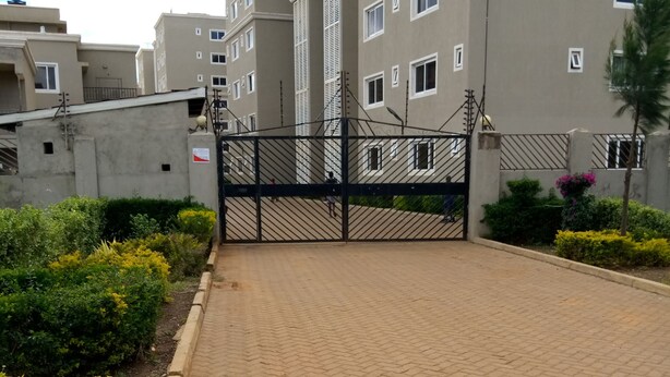 Une entrée fermée par un portail donnant accès à un immeuble résidentiel, avec une allée pavée et des espaces verts aménagés.