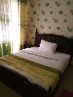 A bed with a dark headboard and white bedding, a green and gold throw blanket, and a floral wallpaper pattern. - Eldoret Kings Square Apartments (Eldoret)