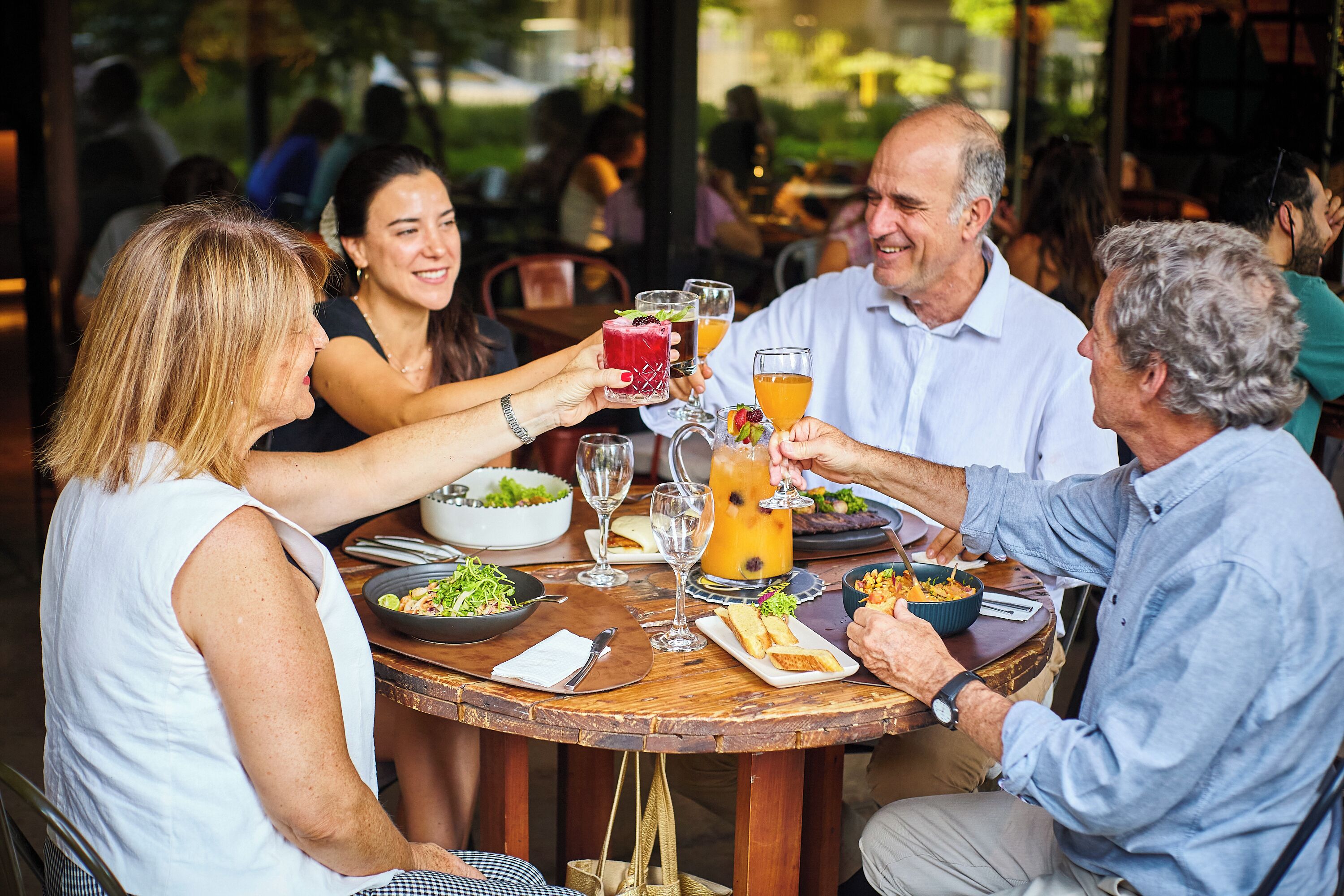 A group of people toasting with drinks at a round wooden table with a bowl of salad and plates of food.