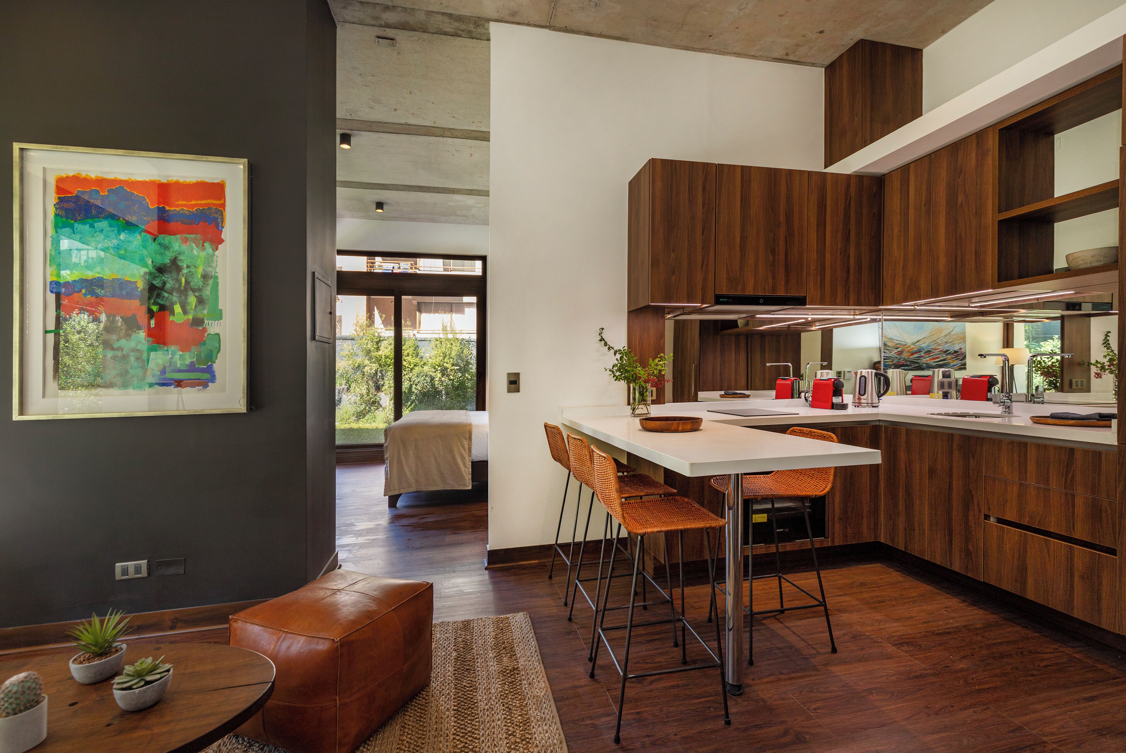 Une cuisine moderne avec des meubles en bois, un îlot central blanc et des tabourets de bar. Un tableau orne le mur et une vue sur l’extérieur est offerte.