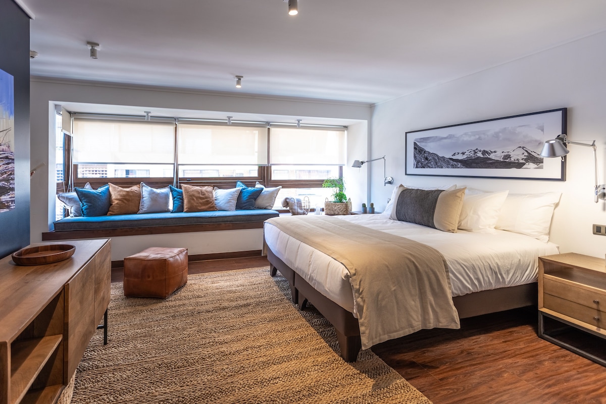 A bedroom with a large bed, a window seat with cushions, a wooden dresser, and a framed picture on the wall.