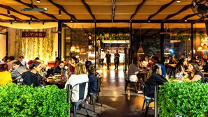 Uma área de refeições interna com mesas e cadeiras, um bar ao fundo e um letreiro com a palavra 'LIVING'.