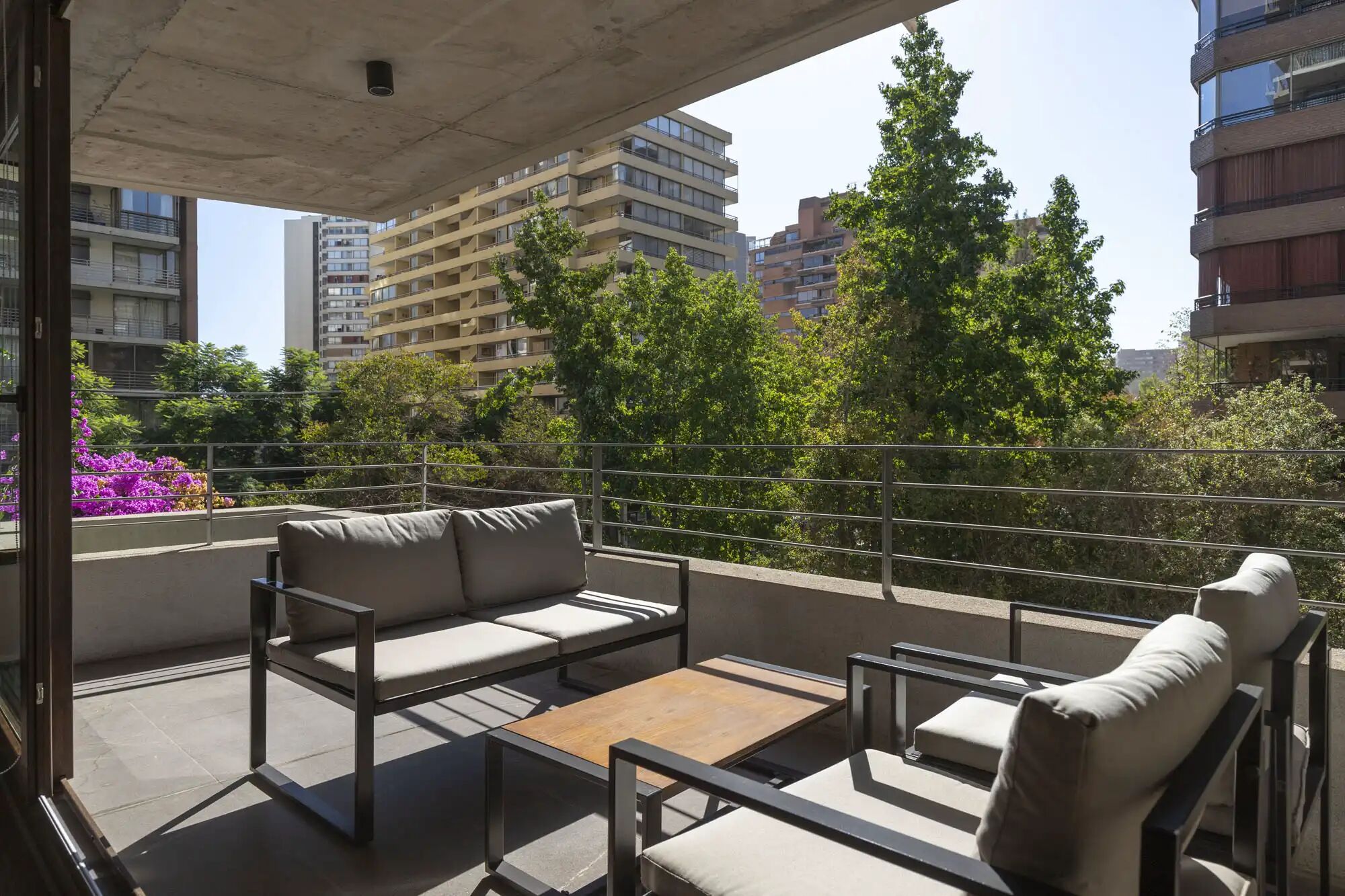 Un balcón con un sofá, una mesita y una silla. Se ven árboles y edificios al fondo.