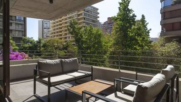 Un balcón con un sofá, una mesita y una silla. Se ven árboles y edificios al fondo.