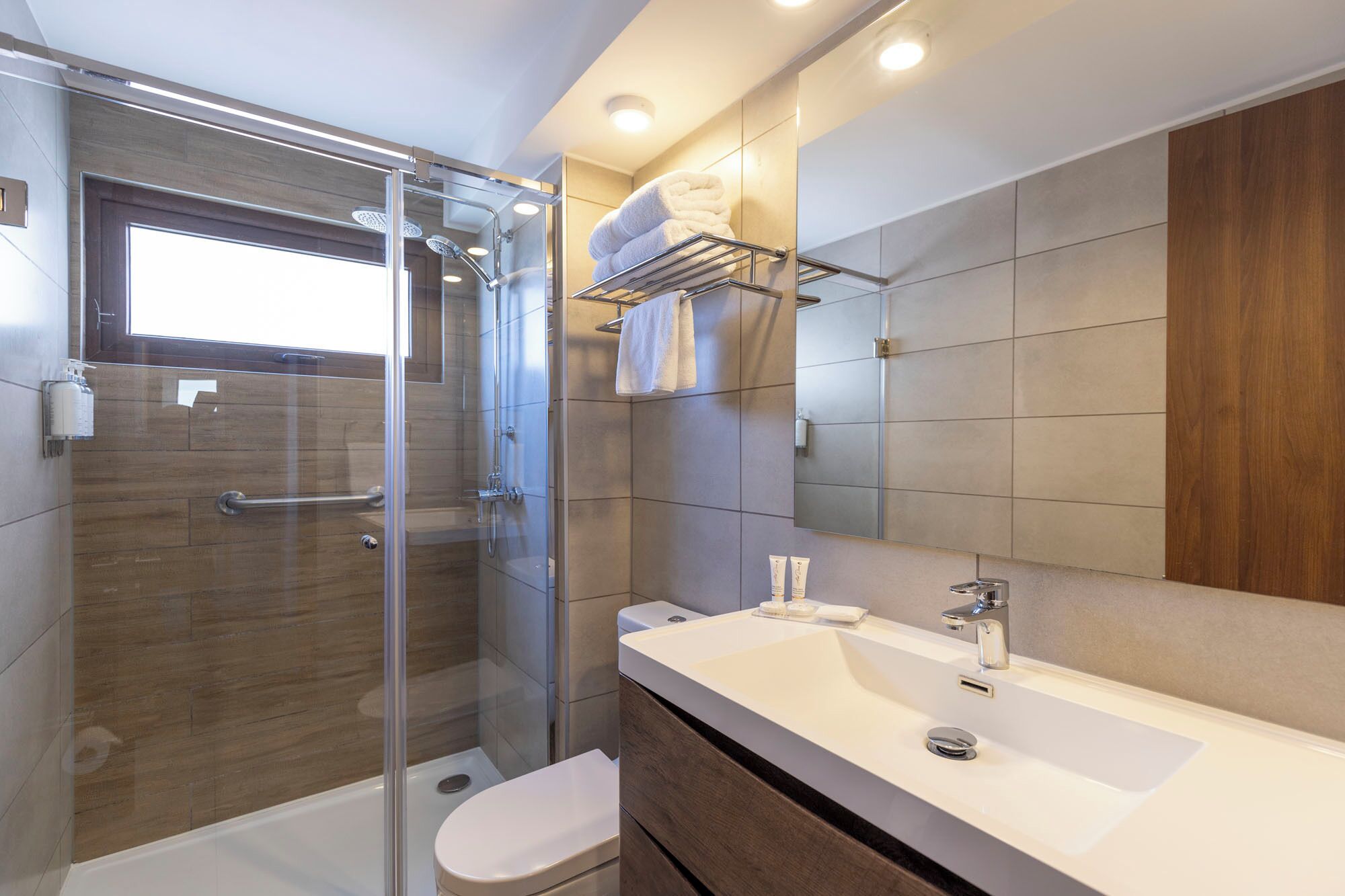 Un baño moderno con ducha envuelta en cristal, un lavabo blanco y un mueble de tocador de madera.