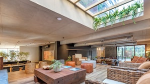 Ampla sala de estar moderna, com teto alto, grandes janelas e diversas opções de assentos, incluindo móveis de vime e uma mesa de centro de madeira.