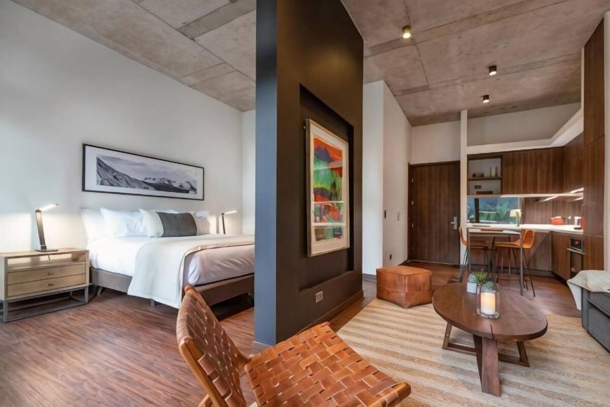 A modern hotel room with a bed, a dining table, a chair, and a kitchen area.