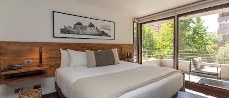 Un dormitorio con una cama grande, cabecera de madera, mesita de noche, teléfono, lámpara, un cuadro enmarcado en la pared y un balcón con una silla.
