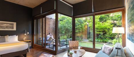 A modern hotel room with a large bed, a sofa, a coffee table, and a view of a lush garden through large windows.